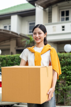 Happy young woman holding moving box and house keys in front of new home. Moving day, real estate purchase, mortgage loan, home ownership and property investment concept.