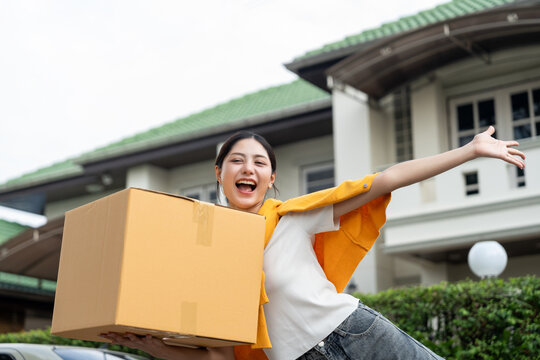Happy young woman holding moving box and house keys in front of new home. Moving day, real estate purchase, mortgage loan, home ownership and property investment concept.