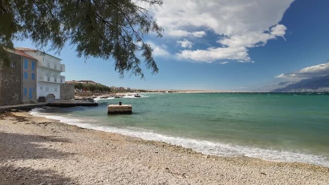 Strong Bura wind at Razanac beach creates bright waves along the empty shore, with stone houses, a long protective pier, and one tiny boat sheltered in the calm pocket.