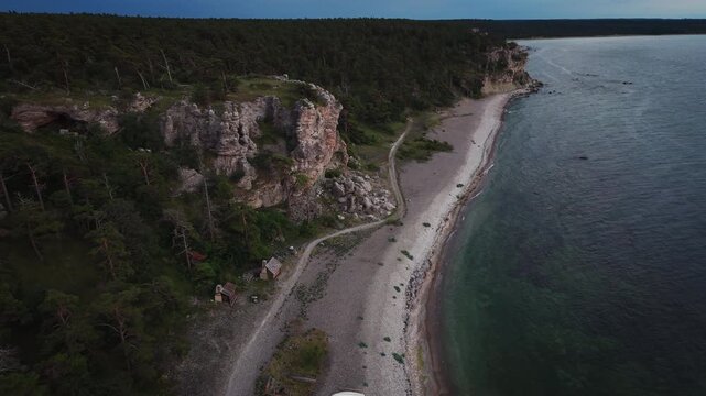Aerial view of forest cliffs and traditional fishing huts beside a rocky beach in Sweden, surrounded by rugged Baltic coastal scenery.
