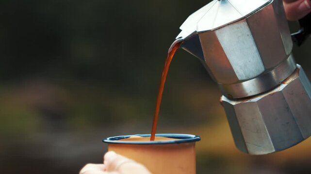 Coffee is being carefully poured into an enamel mug from a steel moka pot held by human hands, showcasing a serene moment of refreshment amidst a quiet and natural setting away from the busy world.
