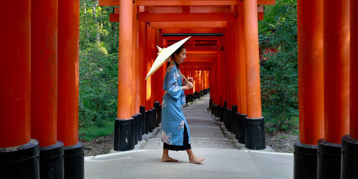 Side View of Woman in Blue Kimono Holding Paper Umbrella Walking Barefoot Through Red Torii Tunnel at Fushimi Inari Kyoto