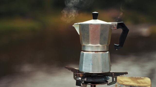 Espresso maker sits on a portable stove with visible steam rising against a soft natural background to represent the perfect way to enjoy a hot coffee beverage while camping deep in the wild forest.