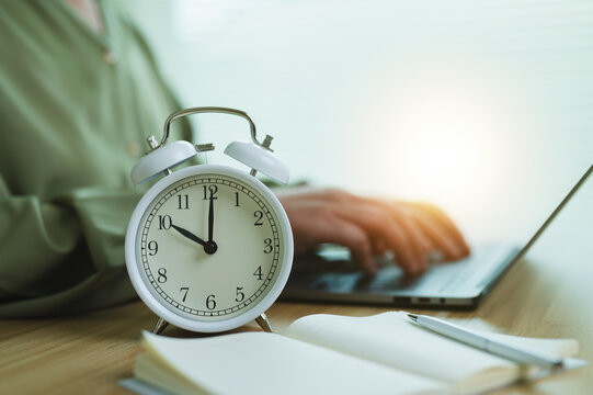 Working against clock, alarm clock on desk with person typing on laptop, concept of deadline, working hours, and morning routine.