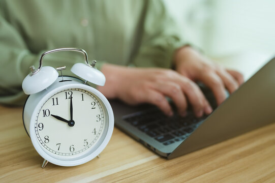 Working against clock, alarm clock on desk with person typing on laptop, concept of deadline, working hours, and daily routine.
