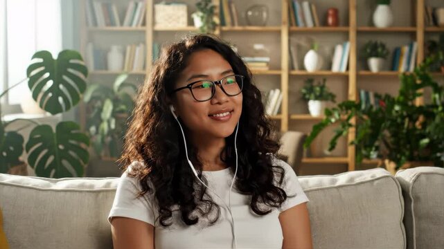 A young adult Asian woman with closed eyes smiles while listening to music on a couch