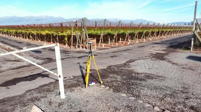 Orbit shot around a GNSS surveying tripod on a rural road flanked by autumn vineyard rows with a vast Andean mountain range stretching across the horizon