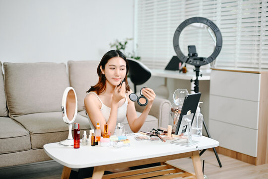 Asian beauty influencer filming a makeup tutorial using cosmetics, skincare products, smartphone and ring light in a modern home studio for social media content.