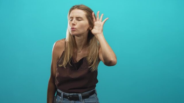 Woman cups hand to ear listening intently in studio with turquoise backdrop, wearing brown camisole and jeans; curiosity focus.