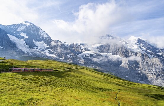 Jungfrau Railway to Jungfraujoch runs through green meadows, above the Kleine Scheidegg stop, rack railway, in the back mountains with peaks Eiger, M&ouml;nch and Jungfrau, Jungfrau region, Bernese Oberland, Switzerland