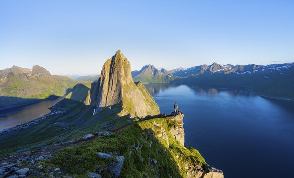 Young man in front of impressive landscape, iconic mountain Segla with steeply sloping cliffs, mountain landscape at Medfjorden fjord, evening light at sunset, Senja island, Troms, Norway