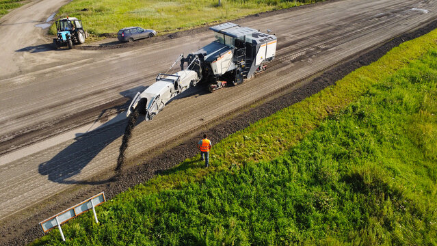 shot captured by drone showing a large road cold recycler machine milling asphalt and discharging reclaimed material along the side of a rural highway.