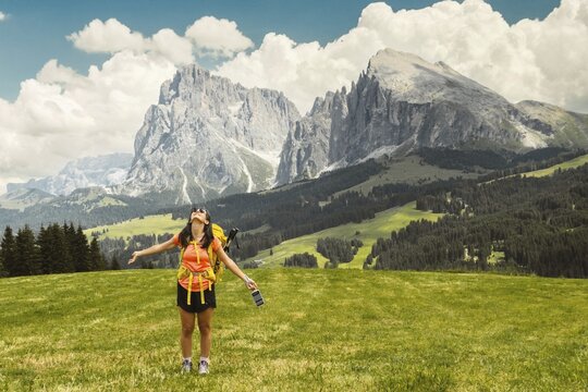 Back view middle aged gray hair woman hiker arms outstretched with yellow backpack overlooking lush green alpine valley misty mountains background Dolomites Alps Italy. Seceda ridge viewpoint