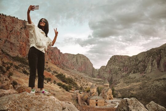 Happy excited female tourist takes a selfie photo against the background of the famous Noravank 13th-century Armenian monastery surrounded by colorful cliffs