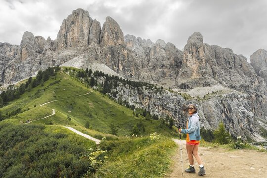 Female hiker with trekking pole turning to look at camera walking winding dirt path along green alpine ridge majestic jagged Cir peaks background Passo Gardena Dolomites Italy