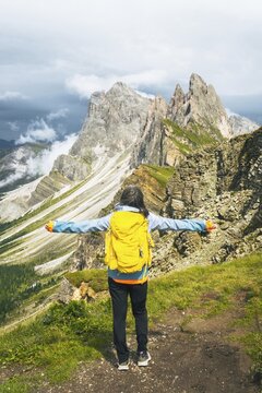 Back view middle aged gray hair woman hiker arms outstretched with yellow backpack overlooking lush green alpine valley misty mountains background Dolomites Alps Italy. Seceda ridge viewpoint