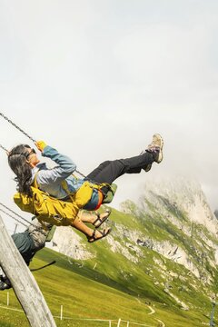 Excited woman with backpack swinging on wooden playground swing lush green hills steep mountain peak misty background Seceda Dolomites Italy summer travel adventure lifestyle