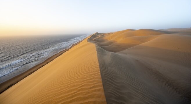 Sand dunes with wave patterns and sea in evening light, wind blowing sand across the dunes, Atlantic coast and sandy desert with sea of dunes, impressive desert landscape at sunset, Sandwich Harbour, Namib Naukluft Park, Namib Desert, Namibia