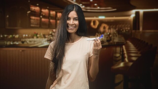 Woman holds toy plane between fingers and smiles, standing by a bar counter with stools inside a building; travel play joy.