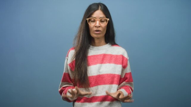 Woman in striped sweater and glasses displays palms forward hand gesture while speaking directly to camera in studio; calm explanation.