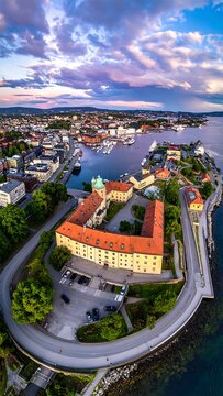 Aerial view of a city by the sea, showcasing a U-shaped building, docks with boats, winding roads, and a colorful sky at dawn