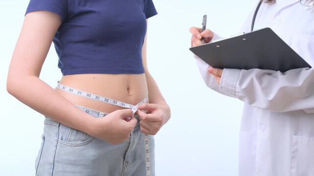 Determined young woman starting weight loss journey measuring waist with tape during medical consultation, professional doctor holding clipboard