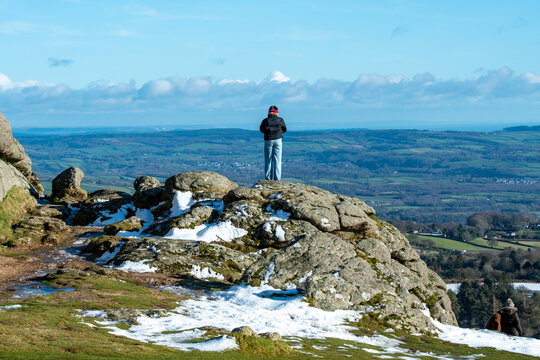 Person stood at the summit of Dartmoor rock looking out over the landscape on a snowy winter's day. Dressed in warm clothing for the climate.