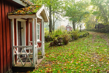 Old red wooden cottage with white trim stands beside a leaf‑covered dirt pat © Lars Johansson