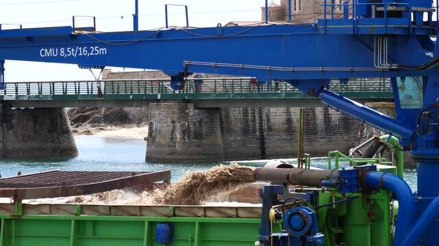 Large blue industrial crane over river. Concrete bridge with pillars visible. Green machinery near water. Mud flows from pipes.