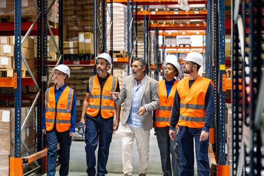 Manager discussing operations with diverse team of industrial workers in safety vests and hard hats inside a busy warehouse