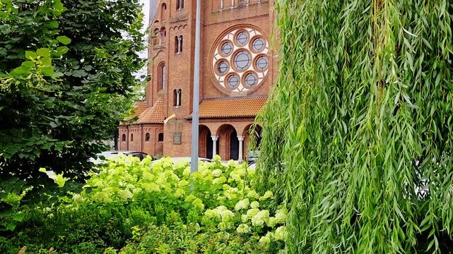 Filmed neogothic red brick church with a round rose window, partially framed by hanging weeping willow branches, green trees, and blooming lime-green hydrangeas in the foreground.