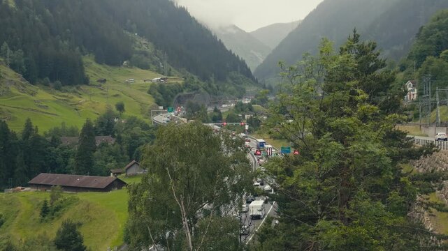 Slow-moving traffic queues on the highway before the Gotthard Tunnel entrance in Switzerland, with spectacular Alpine mountains in the background on a summer day.