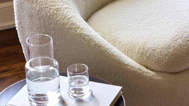 Slow zoom in close-up of glasses of water on a side table beside a cream boucle accent chair in a professionally staged living space