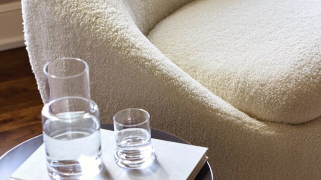 Slow zoom out close-up of glasses of water on a side table beside a cream boucle accent chair in a professionally staged living space.