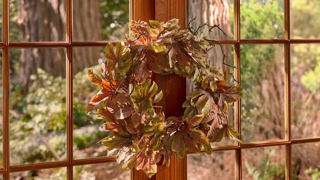 Slow zoom in of a fall leaf wreath viewed from the side on a window with a forest landscape and front yard visible in the background