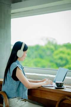 Young Asian woman reading a book with headphones and laptop by the window
