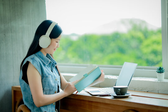 Young Asian woman reading a book with headphones and laptop by the window
