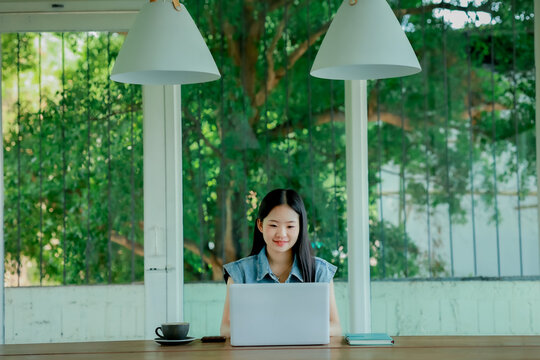 Young Asian woman reading a book with headphones and laptop by the window
