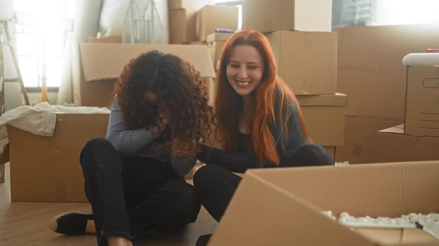 Two women laughing and smiling while unpacking cardboard boxes amid packed cartons in an apartment building; friendship joy.