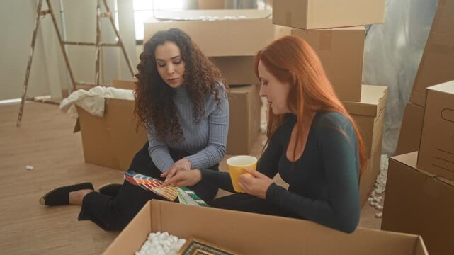 Woman and friend sit on floor holding a yellow coffee mug and examining paint color swatches while surrounded by moving boxes in a building; friendship planning contentment.
