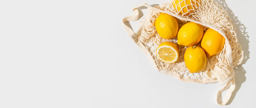 Yellow citrus lemons in reusable mesh bag, fresh fruit and green leaves displayed against light gray background. Eco friendly shopping, zero waste habits and sustainable choices support branding.