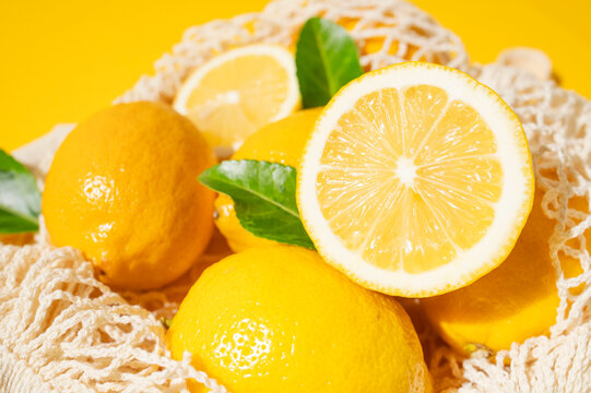 Fresh lemons in cotton mesh bag, whole and sliced citrus with green leaves on vivid yellow background. Juicy fruit, fresh produce and citrus appeal support food marketing, retail, lifestyle.