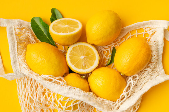 Whole and sliced lemons in reusable mesh bag, bright citrus composition with green leaves on yellow background. Organic produce, natural freshness and sustainable shopping concept suit food campaigns.