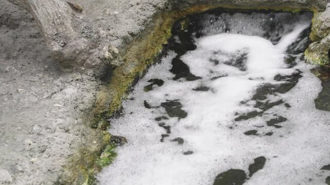 Natural geothermal hot spring pool with bubbling white foam and flowing thermal water. Volcanic mineral deposits on rocky edges with sulfur and algae. Geologic activity in a thermal park area.