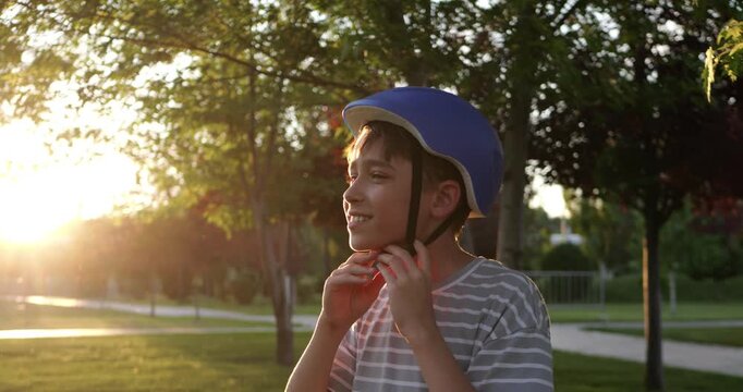 Happy boy wearing helmet adjusts straps in park at sunset. Child prepares for outdoor activity, enjoying warm evening light. Safety first for fun biking adventures.