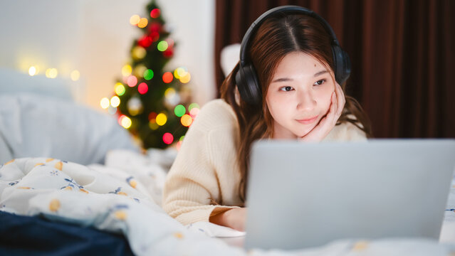 Young adult asian woman using laptop relax sitting in cozy winter bedroom with christmas tree enjoy alone at home for the holidays