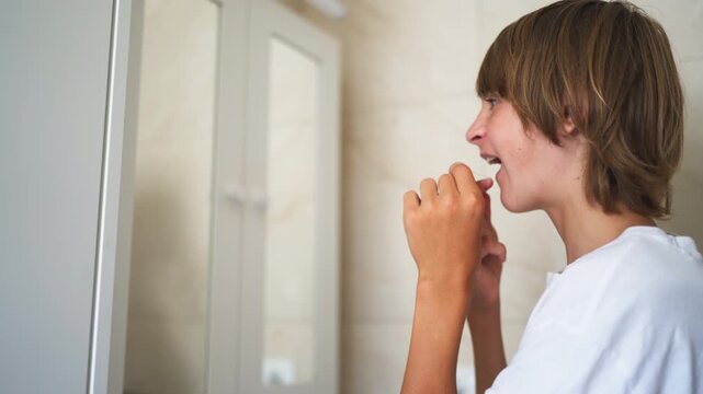 Teenage boy carefully flossing between his teeth while looking in the bathroom mirror, practicing daily oral hygiene and maintaining healthy teeth as part of his personal care routine.