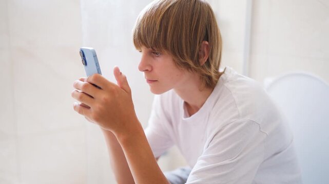 Young boy sitting on the toilet in a bright bathroom, completely absorbed in his smartphone. Concept of digital addiction and excessive screen time in modern youth