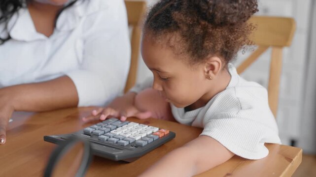 Little black girl studying mathematic, using calculator and pressing buttons. Lesson of addition and subtraction in primary school, homeschooling and family education, mother or teacher teaching