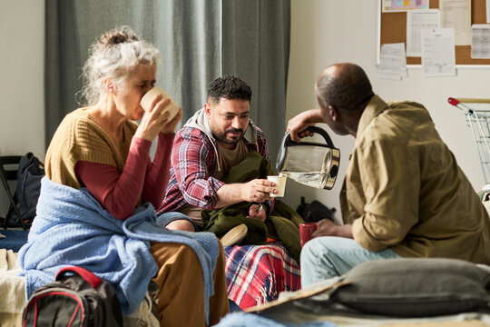 Diverse adults resting and sharing hot drinks in temporary shelter, supporting each other during displacement and crisis. Useful for refugee aid, emergency housing, community support